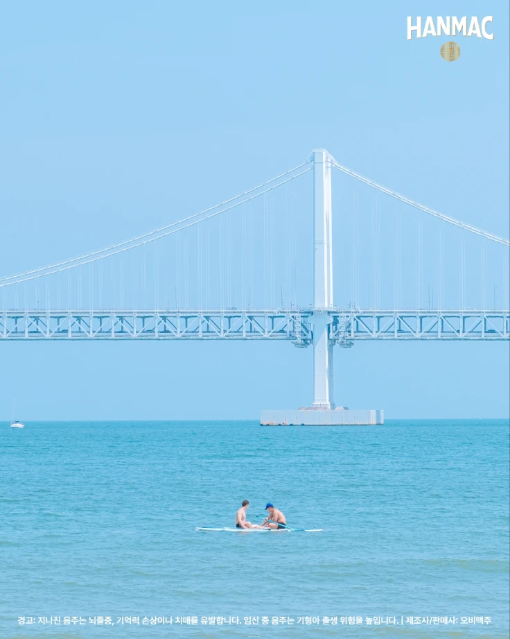 한맥 장소 디스커버리 세번째 에피소드 부드러운 바다와 파도 명소 네번째 부산 광안리 해수욕장 바다 위에 서핑보드를 타는 두 사람과 뒤에 광안대료가 보입니다.